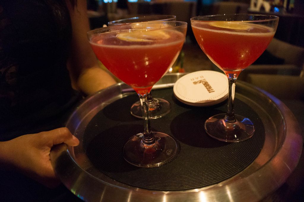 Three colorful drinks in martini glasses on a tray with a stack of coasters that say "L Bar".