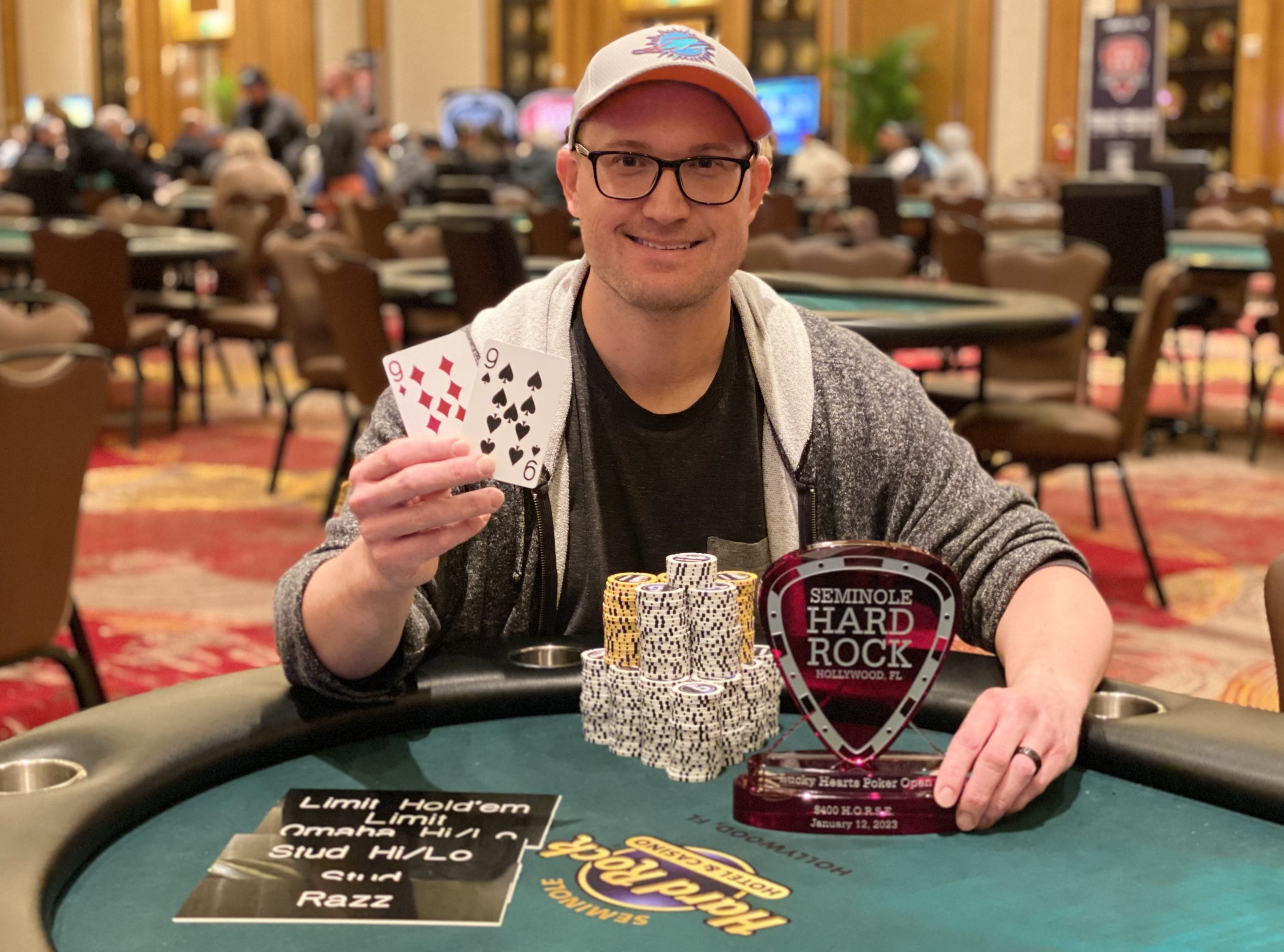 A smiling Mark Hurst holds pocket nines as he poses with the HORSE trophy and a big pile of poker chips.